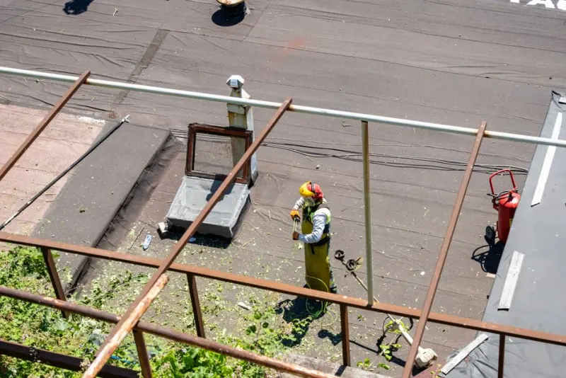 Industrial worker in protective gear trims plants on a rooftop in daylight.